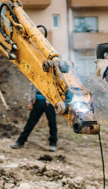 excavator welding on a construction site with sparks flying and worker in background heavy machinery use